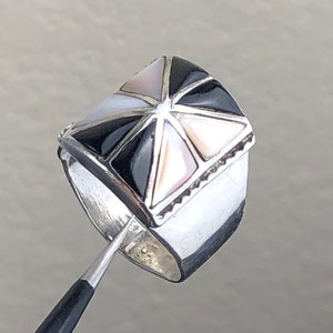 May include: A silver ring with a square-shaped top. The top is inlaid with black and white shell pieces in a geometric pattern.