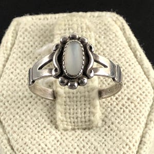 May include: A silver ring with an oval, light-colored stone set in a decorative bezel. The ring has a split shank design and is displayed on a textured, off-white surface. The ring's design includes small, round embellishments around the stone.