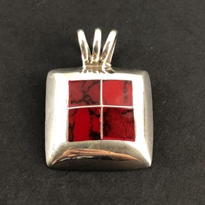 May include: A square silver pendant featuring a central inlay of four red stone segments with dark veining. The pendant has a polished metallic frame and a triple-pronged bail at the top for attaching to a chain or cord, creating a geometric design.