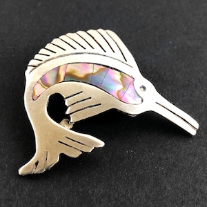 Puede incluir: Broche de pez estilizado en tono plateado con incrustaciones de concha de abulón iridiscente. El broche presenta un diseño detallado con un hocico largo y puntiagudo y una cola curvada. La concha de abulón muestra tonos de rosa, morado y verde.