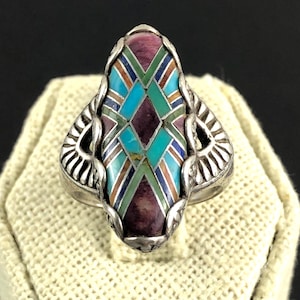 May include: A silver ring with an elongated oval face. The face features a mosaic design with turquoise, purple, green, and brown inlays. The ring has decorative side accents.