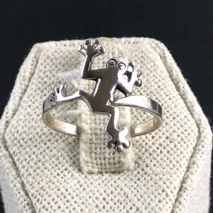 May include: A silver-toned ring featuring a stylized gecko design. The gecko is depicted with outstretched limbs and a textured body, set against a simple band. The ring is displayed on a textured, off-white surface.
