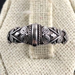 Peut inclure: Une bague en argent avec un design orné de volutes, d'éléments floraux et d'une barre rectangulaire centrale. La bague a un aspect détaillé et texturé, mettant en valeur un savoir-faire complexe. La bague est présentée sur un tissu beige.
