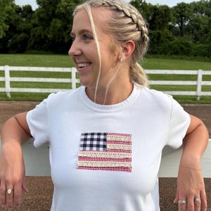 Puede incluir: Camiseta blanca con un diseño de bandera americana cosido. La bandera presenta un cuadrado de cuadros azul marino y blanco, rayas de cuadros rojos y blancos y detalles de encaje color crema. La camiseta tiene cuello redondo y mangas cortas.