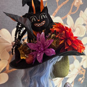 May include: A black witch hat adorned with a plush black cat face, orange and purple artificial flowers, gold-toned roses, and a small red pumpkin. The hat features a pointed crown and a wide brim, decorated with dark foliage and orange accents.