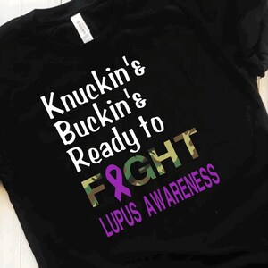 May include: Black t-shirt with a camouflage pattern and purple ribbon. The text on the shirt reads "Knuckin' Buckin' & Ready to Fight Lupus Awareness".