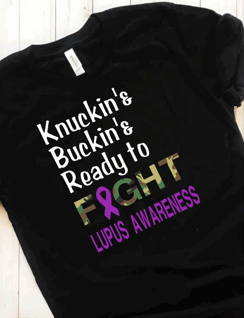 May include: Black t-shirt with a camouflage pattern and purple ribbon. The text on the shirt reads "Knuckin' Buckin' & Ready to Fight Lupus Awareness".