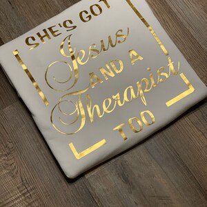 May include: White t-shirt with gold lettering that reads "She's got Jesus and a therapist too."