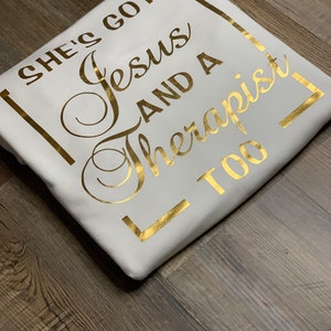May include: White t-shirt with gold foil lettering that reads "She's got Jesus and a therapist too" in a square frame.