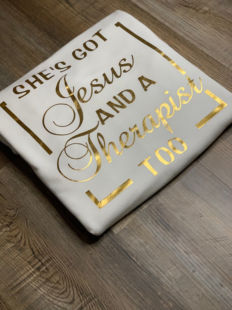 May include: White t-shirt with gold foil lettering that reads "She's got Jesus and a therapist too" in a square frame.