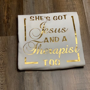 May include: White t-shirt with gold lettering that reads "She's got Jesus and a therapist too."