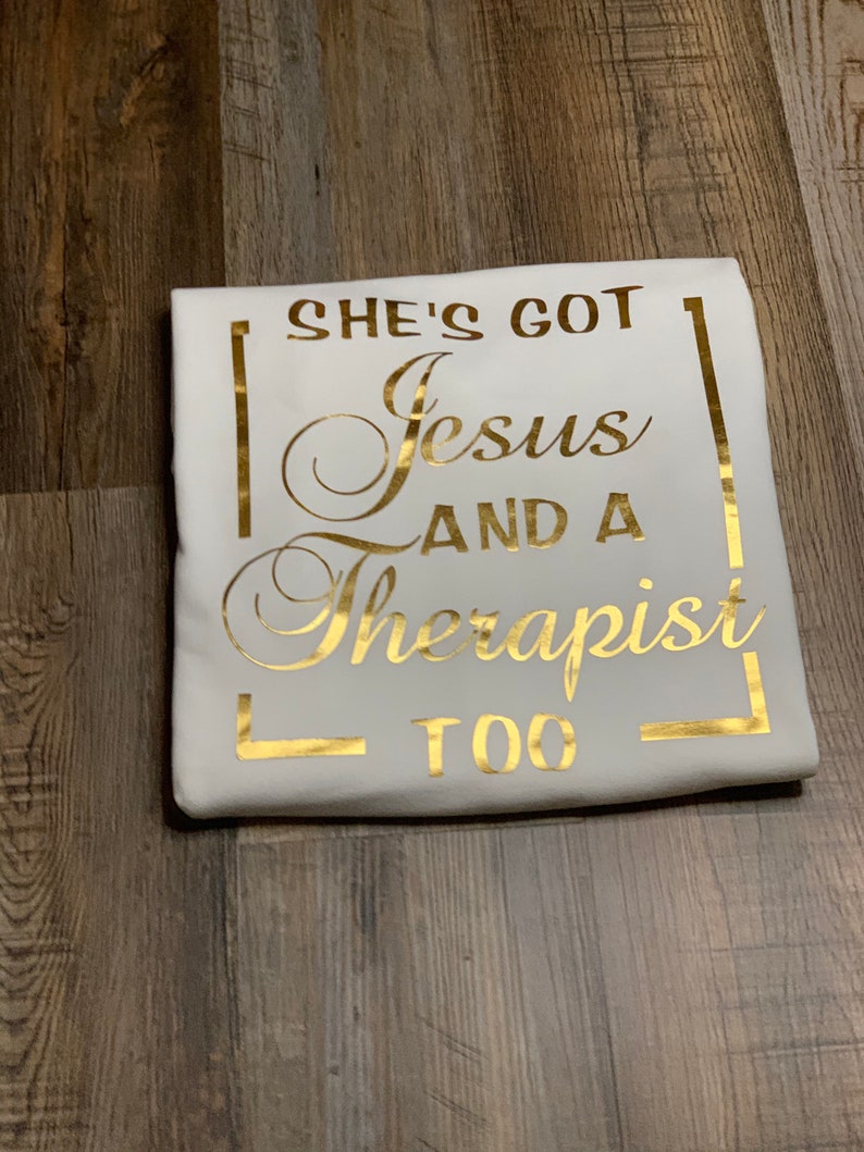 May include: White t-shirt with gold lettering that reads "She's got Jesus and a therapist too."