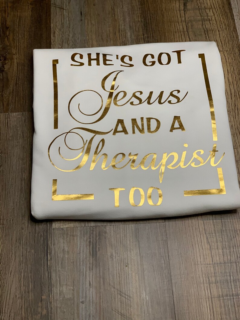 May include: White t-shirt with gold foil lettering that reads "She's got Jesus and a therapist too."