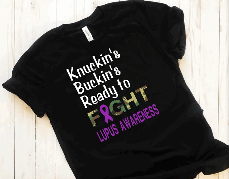 May include: Black t-shirt with a white graphic that says "Knuckin' & Buckin' & Ready to Fight" with a purple ribbon and camouflage pattern. The text "Lupus Awareness" is below the graphic.