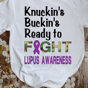 May include: White t-shirt with the text "Knuckin' & Buckin' & Ready to FIGHT" in black. The word "FIGHT" is in camouflage print with a purple ribbon in the middle. The text "LUPUS AWARENESS" is in purple below the word "FIGHT".