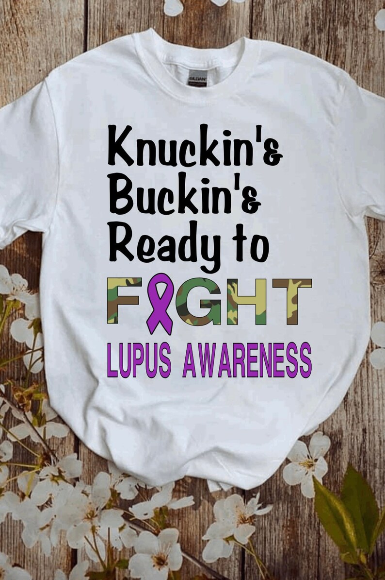 May include: White t-shirt with the text "Knuckin' & Buckin' & Ready to FIGHT" in black. The word "FIGHT" is in camouflage print with a purple ribbon in the middle. The text "LUPUS AWARENESS" is in purple below the word "FIGHT".