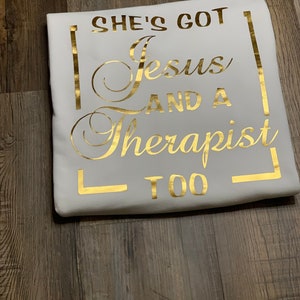 May include: White t-shirt with gold lettering that reads "She's got Jesus and a therapist too."