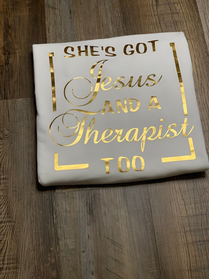 May include: White t-shirt with gold lettering that reads "She's got Jesus and a therapist too."