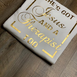 May include: White t-shirt with gold lettering that reads "She's got Jesus and a therapist too" in a square frame.