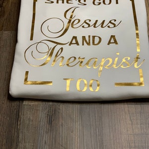 May include: White t-shirt with gold lettering that reads "She's got Jesus and a Therapist too".