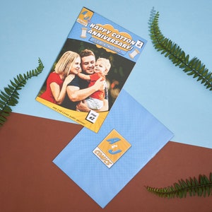 May include: A comic book cover with a photo of a family of three. The cover is yellow and blue with the text "Happy Cotton Anniversary! It's all for you..." and "Issue #2". The comic book is on a blue surface with a blue envelope with a yellow and blue logo that says "A&J Comics".