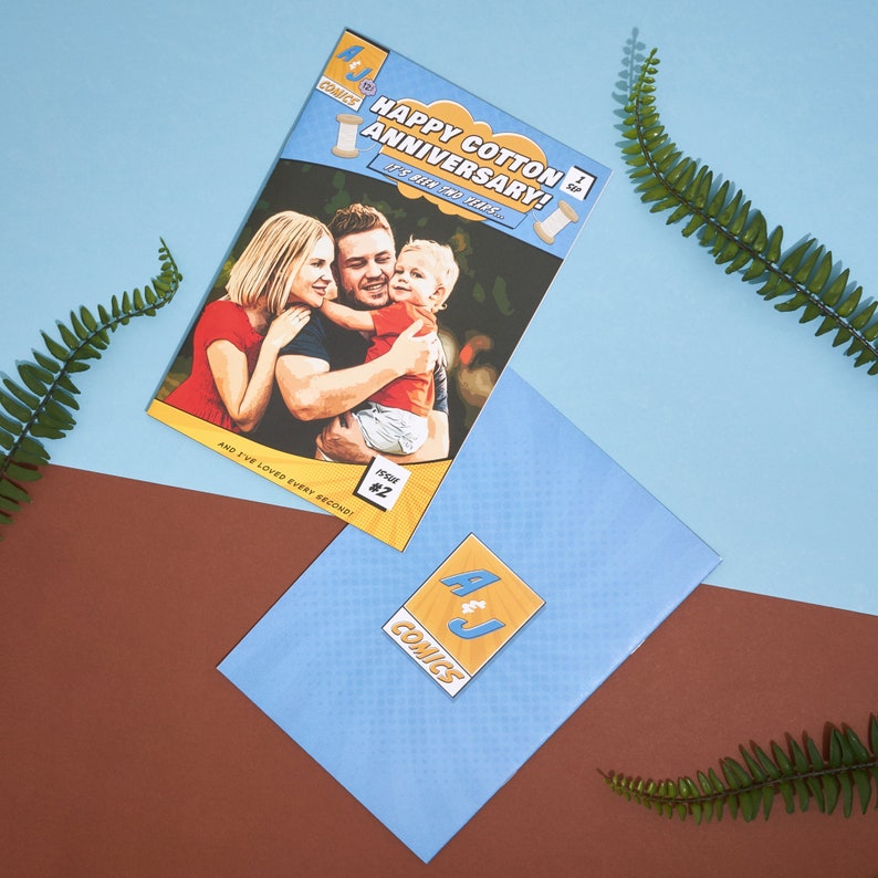 May include: A comic book cover with a photo of a family of three. The cover is yellow and blue with the text "Happy Cotton Anniversary! It's all for you..." and "Issue #2". The comic book is on a blue surface with a blue envelope with a yellow and blue logo that says "A&J Comics".