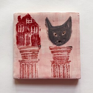 May include: A ceramic tile with a pink background and two red columns. The columns have a red house and a black cat head on top.
