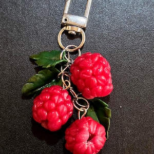 May include: A keychain with three vibrant red raspberry charms and green leaves. The charms are attached to a silver-coloured clasp. The raspberries have a textured surface, and the leaves are detailed.