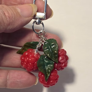 May include: A silver keychain with a cluster of three red raspberry charms and green leaves. The raspberries have a textured surface, and the leaves have detailed veining. The keychain is attached to a silver clasp.