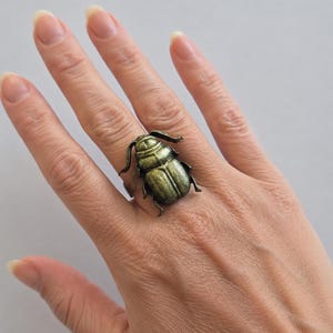 Könnte beinhalten: Ein bronzefarbener Ring mit einem detaillierten Käfer-Design. Der Ring wird an einem Finger getragen und zeigt den strukturierten Körper und die Fühler des Insekts. Die metallische Oberfläche verleiht dem Ring ein Vintage- oder Antik-Aussehen.