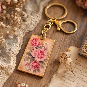 May include: A rectangular keyring featuring a vintage-style illustration of three pink roses with green leaves on a beige background. The pendant is attached to a gold-toned metal split ring and a lobster clasp connector via a short chain.