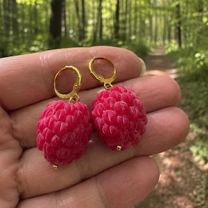 Op de afbeelding: Een paar hangende oorbellen met realistische, getextureerde, felroze framboosvormige bedels die aan kleine, ronde goudkleurige metalen ringetjes hangen. Elke framboosbedel heeft een gedetailleerd, bobbelig oppervlak en een klein gouden kraaltje.