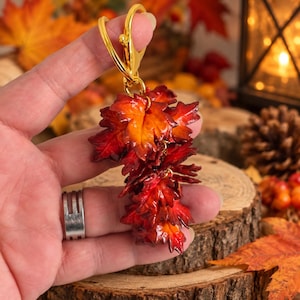 Può includere: Un portachiavi in metallo dorato con un grappolo di piccole foglie d'acero realistiche in vivaci tonalità di rosso, arancione e giallo. Le foglie sono sovrapposte e fissate a una robusta chiusura a moschettone dorata, creando un ciondolo autunnale.