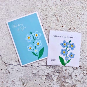 May include: Two greeting cards featuring blue forget-me-not flowers. The card on the left reads "Thinking of You". The card on the right reads "Forget-Me-Not" and "Border in a Box".