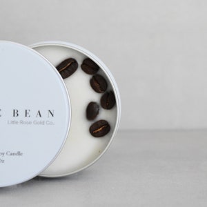 May include: A white soy candle in a tin container with the text "The Bean Little Rose Gold Co." and coffee beans on top of the wax.
