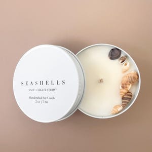 May include: A white round metal tin containing a soy candle with a single wick. The lid is placed beside the candle and features the text SEASHELLS SALT + LIGHT STORY Handcrafted Soy Candle 2 oz | 7 hrs. Several small seashells are embedded in the wax.
