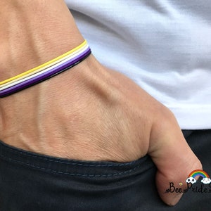 May include: A non-binary pride bracelet with yellow, white, purple, and black bands. The bracelet is on a wrist, with a white top and dark trousers in the background. The image includes the text "Bee Pride Style".
