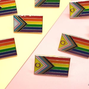 May include: Eight enamel pins featuring the Progress Pride flag. The pins are arranged on a yellow and pink background. The pins are all the same size and shape, and they are all in focus. The pins are a great way to show your pride and support for the LGBTQ+ community.