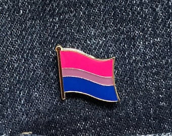 May include: A gold-colored pin with a bisexual pride flag design. The flag has pink, light blue, and dark blue stripes. The pin is on a blue denim background.