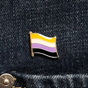 May include: A gold-coloured pin with a yellow, white, purple and black striped flag design. The pin is on a blue denim background.