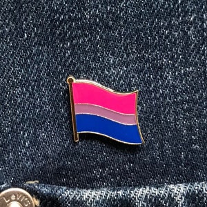 May include: A gold-coloured pin featuring a bisexual pride flag design. The flag has pink, light blue and dark blue stripes. The pin is on a blue denim background.