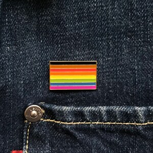 May include: A rectangular enamel pin featuring a rainbow flag design with black, brown, orange, red, yellow, green, blue, and pink stripes. The pin is set against a dark blue denim background.