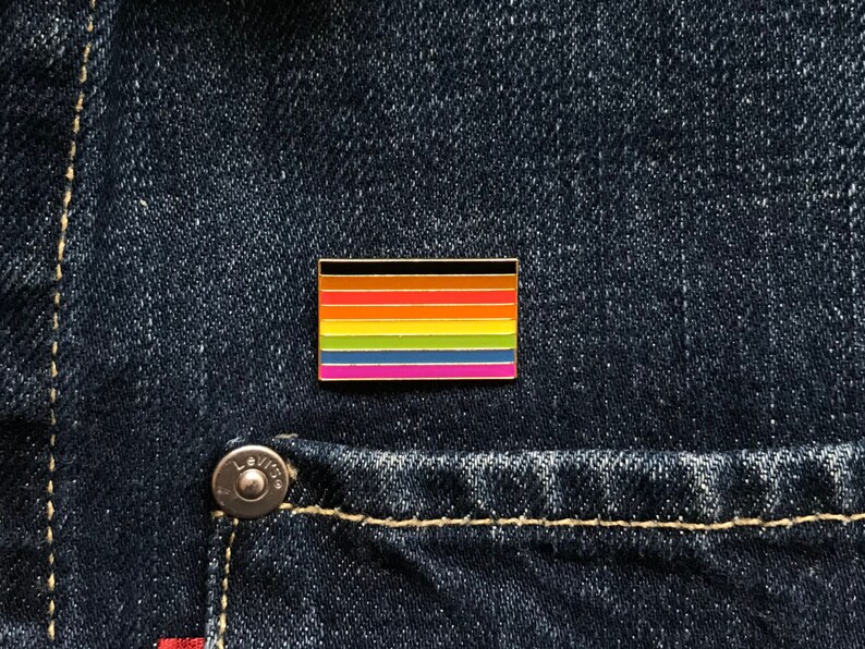 May include: A rectangular enamel pin featuring a rainbow flag design with black, brown, orange, red, yellow, green, blue, and pink stripes. The pin is set against a dark blue denim background.