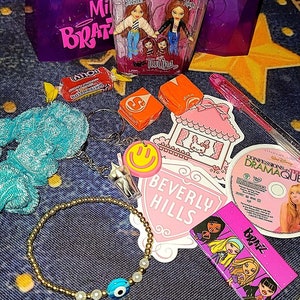 Può includere: Una collezione di articoli a tema Bratz che include un contenitore di plastica rosa e viola con le parole "Mini Bratz" su di esso, un contenitore di plastica trasparente con due bambole Bratz all'interno, un adesivo rosa e bianco con le parole "Beverly Hills" su di esso, un adesivo rosa e bianco con una casa su di esso, un giocattolo di peluche blu e bianco, un braccialetto d'oro con un ciondolo di occhio blu, una carta rosa e viola con le parole "Bratz" su di essa, un CD con le parole "Confessions of a Teenage Drama Queen" su di esso, e un involucro di caramelle rosa e bianco.