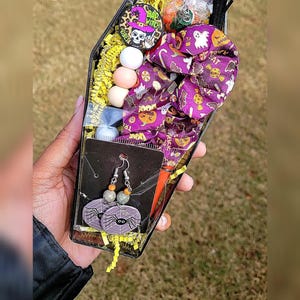 Peut inclure: Une boîte noire en forme de cercueil remplie d'articles sur le thème d'Halloween, dont un chouchou à cheveux à motifs violets et blancs, un ensemble de boucles d'oreilles en toile d'araignée, un petit sac de bonbons et un bouton rond avec le texte "Bad Witch Vibes".