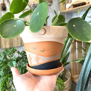 May include: A terracotta planter with a desert scene painted on it. The scene includes a crescent moon, cacti, and birds flying across a brown and beige landscape. The planter is sitting on a saucer and is filled with a green plant.