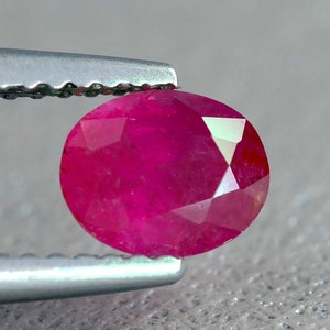 May include: A vibrant, deep red ruby gemstone with a faceted cut, held by silver tweezers. The ruby is oval-shaped and has a high polish.