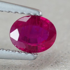 May include: A vibrant, oval-shaped ruby gemstone with a deep red hue. The gemstone is held in place by a pair of tweezers.