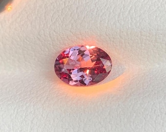 May include: An oval-cut gemstone with a mix of pink and red hues. The gem has a faceted surface that reflects light, creating a sparkling effect. The gemstone is set against a white background.