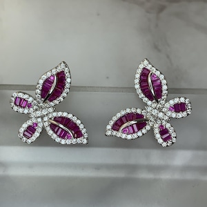 May include: A pair of silver-toned earrings in a floral design. Each earring features a cluster of magenta-colored rectangular stones surrounded by a border of small, clear, round stones. The earrings are set against a light background.